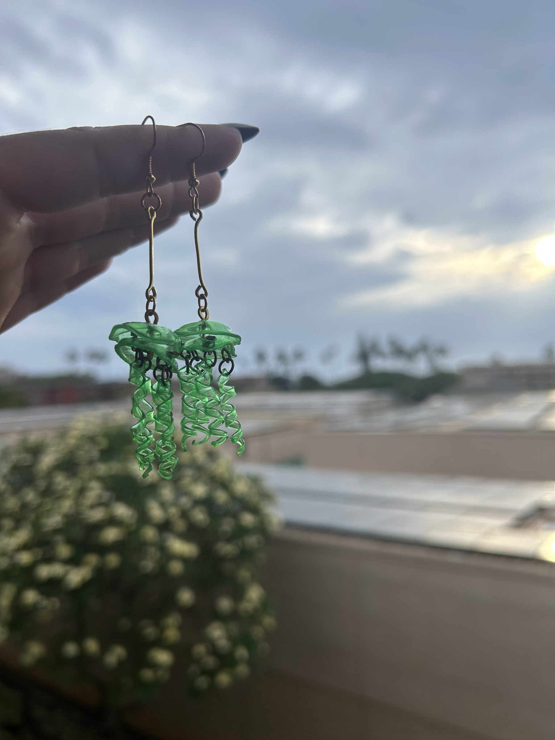 Green jellyfish earrings held against a soft Maui sky.